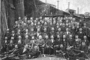 Pracownicy warsztatów na tle dużej parowozowni (towarowej). Zdjęcie z 1899 roku łódzkiego fotografa A. Kuliga