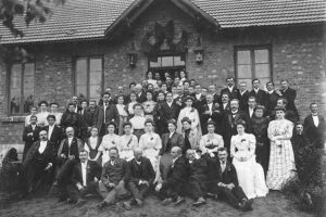Grupa pracowników kolei z rodzinami przed wejściem do szpitalika kolejowego zbudowanego na początku XX wieku przy przejeździe kolejowym. Zdjęcie wykonane przez zakład fotograficzny Stanisławy Odembskiej w Kamiennej